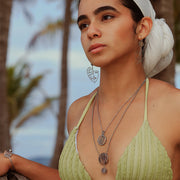 A woman in a light green bikini top and white hair scarf wearing two necklaces, a bracelet and Into The Jungle earrings in front of palm trees.  