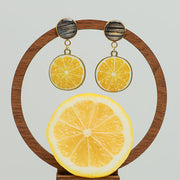 Limoncello Earrings on a circular wooden stand on a white background with a half of a lemon at the bottom of the image.