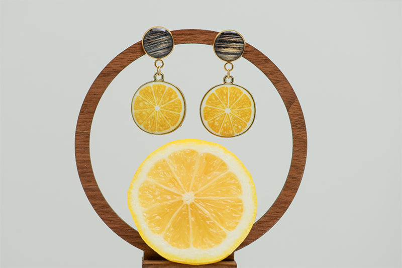 Limoncello Earrings on a circular wooden stand on a white background with a half of a lemon at the bottom of the image.