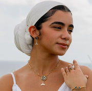 A woman in a white dress and white hair scarf wearing a bracelet, a ring, a necklace and pair of earrings standing in front of a grassy area. 