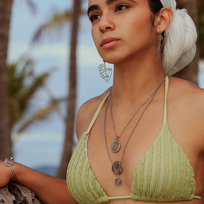 A woman in a light green bikini top and white hair scarf wearing two necklaces, a bracelet and earrings in front of palm trees. 