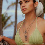 A woman in a light green bikini top and white hair scarf wearing two necklaces, a bracelet and earrings in front of palm trees. 