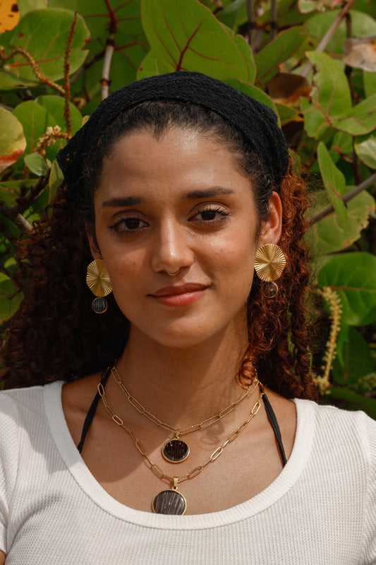 A woman in a white t-shirt wearing two necklaces and a pair of earrings in front of a tree. 