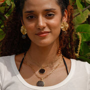 A woman in a white t-shirt wearing two necklaces and a pair of earrings in front of a tree. 