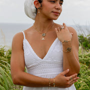 A woman in a white dress and white hair scarf wearing four bracelets, three rings, a necklace and pair of earrings standing in front of a grassy area. 
