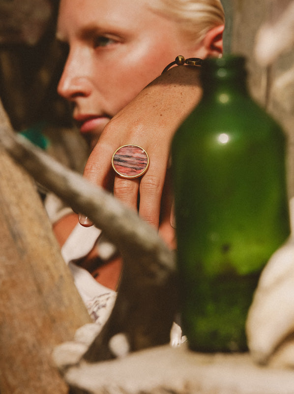  A woman's hand and face behind a glass green bottle and driftwood wearing a Gold Statement Ring.