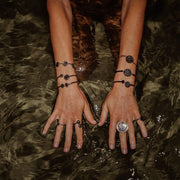 A person's Hands and arms showing four rings, six bracelets over a dark shallow watery background. 