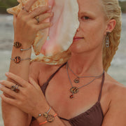 A woman in a brown bikini top in front of the ocean holding a conch shell and wearing three rings, four bracelets, two necklaces and earrings. 