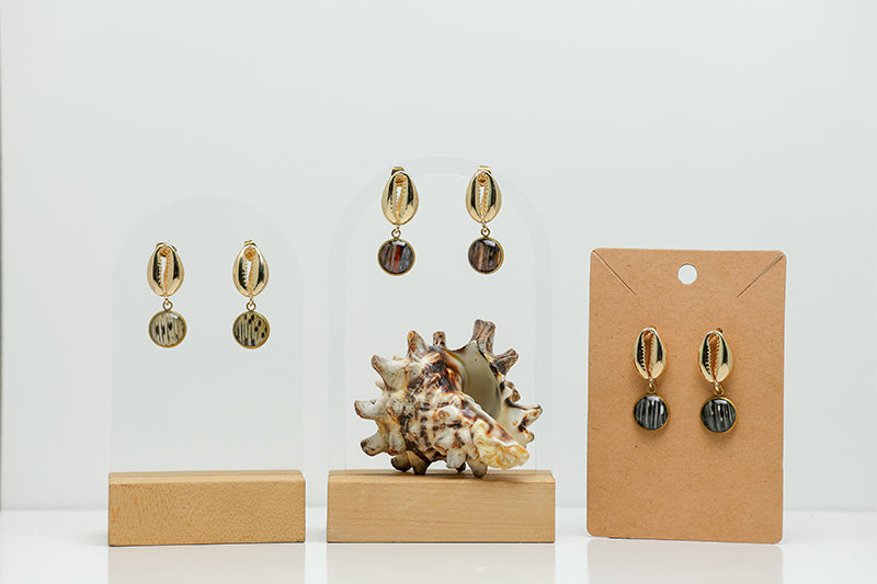 Three pairs of Gold Cowrie Shell Posts on stands on a white background with a shell at the bottom of the image. 