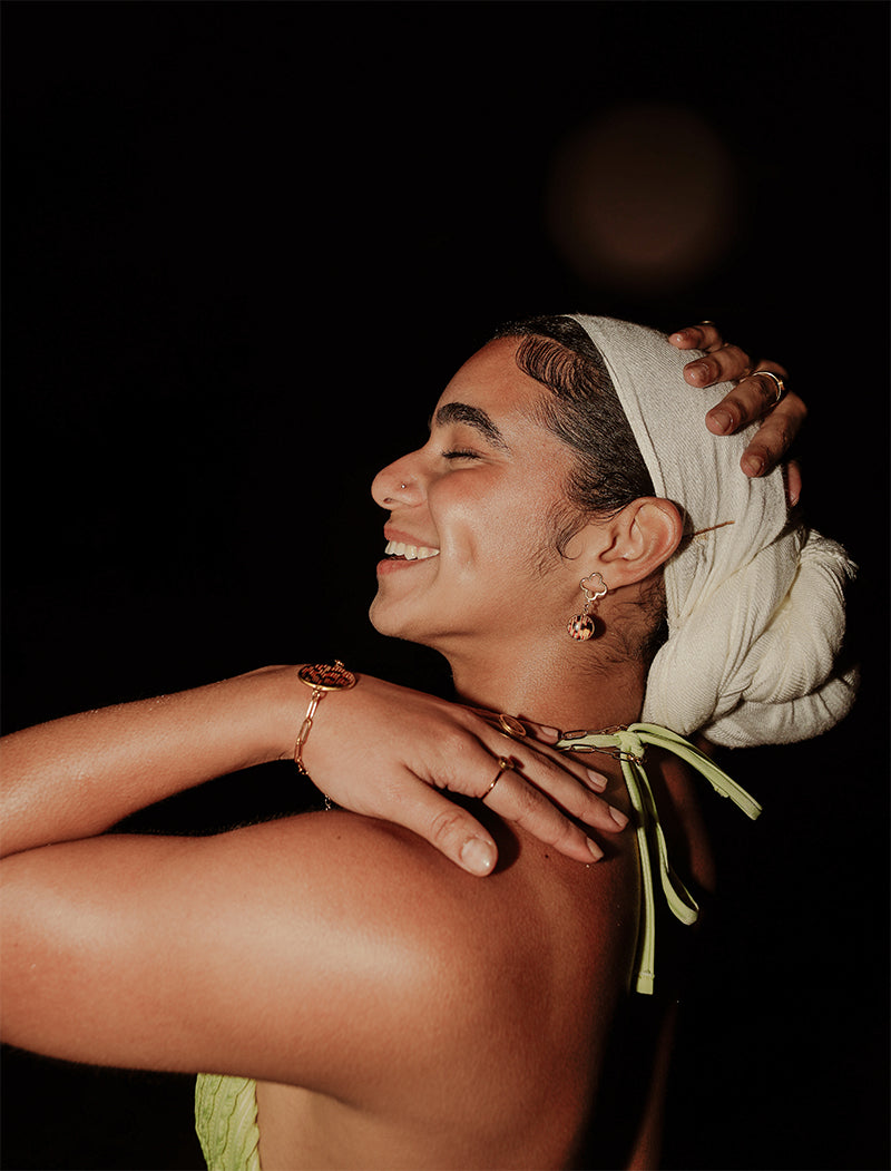 A woman in a light green bikini top and white hair scarf wearing three rings, a bracelet and earrings on a dark background.