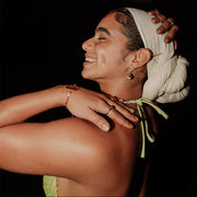A woman in a light green bikini top and white hair scarf wearing three rings, a bracelet and earrings on a dark background.