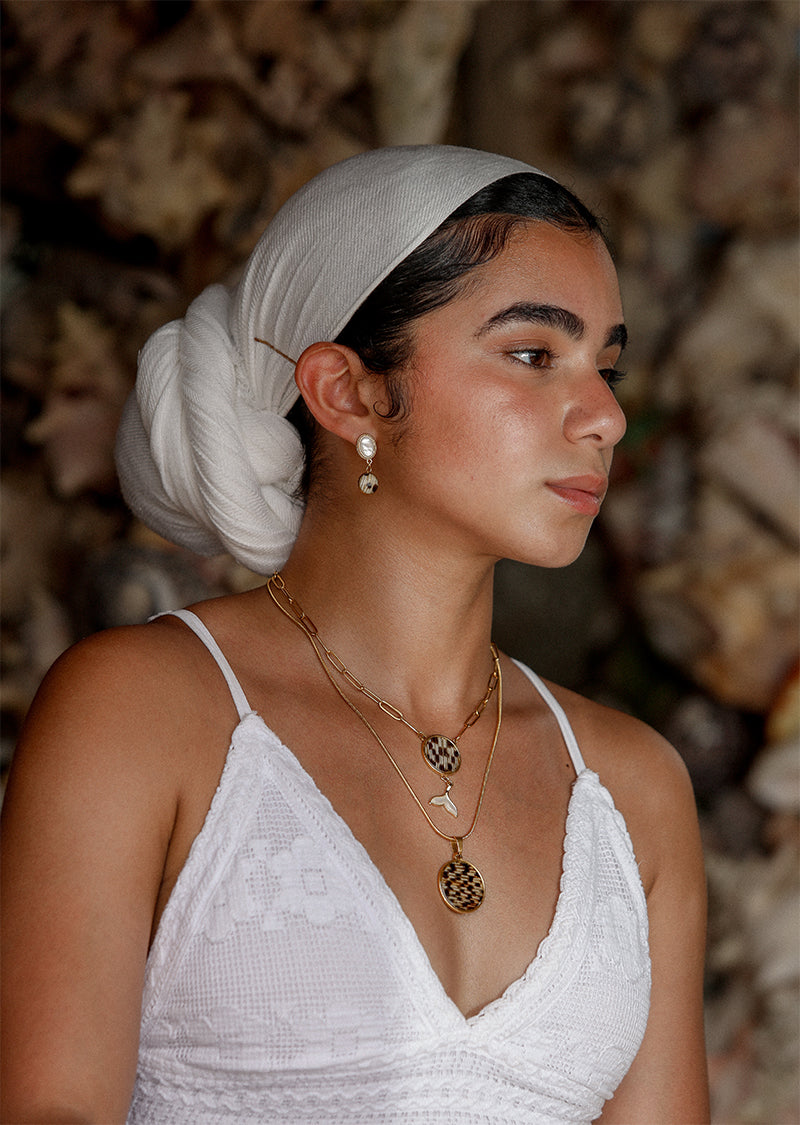 A woman in a white dress and white hair scarf wearing Freshwater Pearl Earrings and two necklaces in front of a dark rocky background.