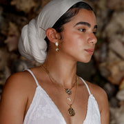 A woman in a white dress and white hair scarf wearing Freshwater Pearl Earrings and two necklaces in front of a dark rocky background.