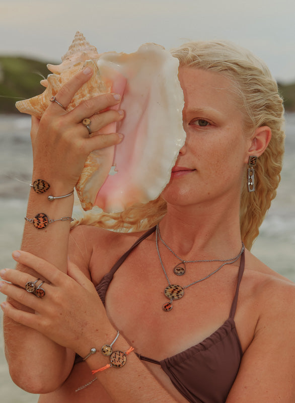 A woman in a brown bikini top in front of the ocean holding a conch shell wearing three rings, four bracelets, two necklaces and earrings. 
