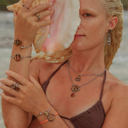 A woman in a brown bikini top in front of the ocean holding a conch shell wearing three rings, four bracelets, two necklaces and earrings. 