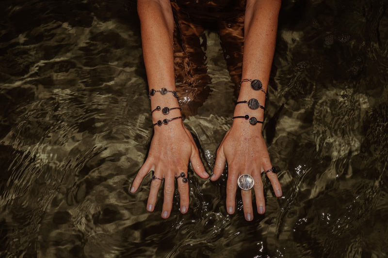 Model's hands with four rings and six bracelets on a dark shallow watery background. 