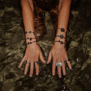 Model's hands with four rings and six bracelets on a dark shallow watery background. 