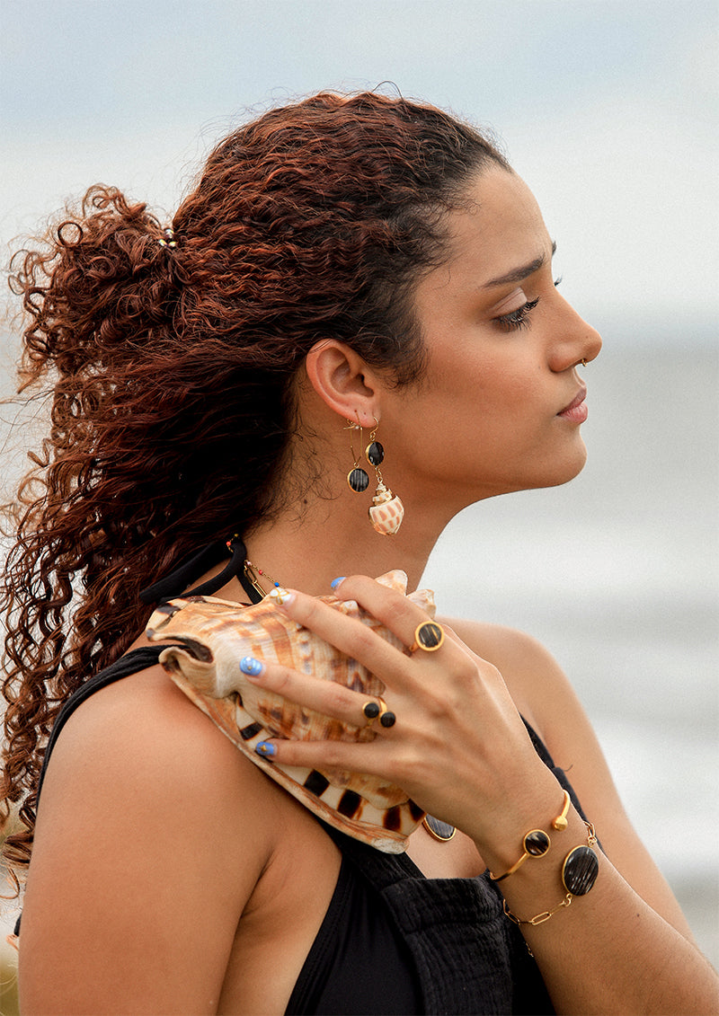 A woman holding a conch shell in. ablack top wearing two bracelets, two rings, two earrings.  