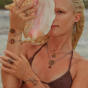 A woman in a brown bikini top in front of the ocean holding a conch shell wearing three rings, four bracelets, two necklaces and earrings. 