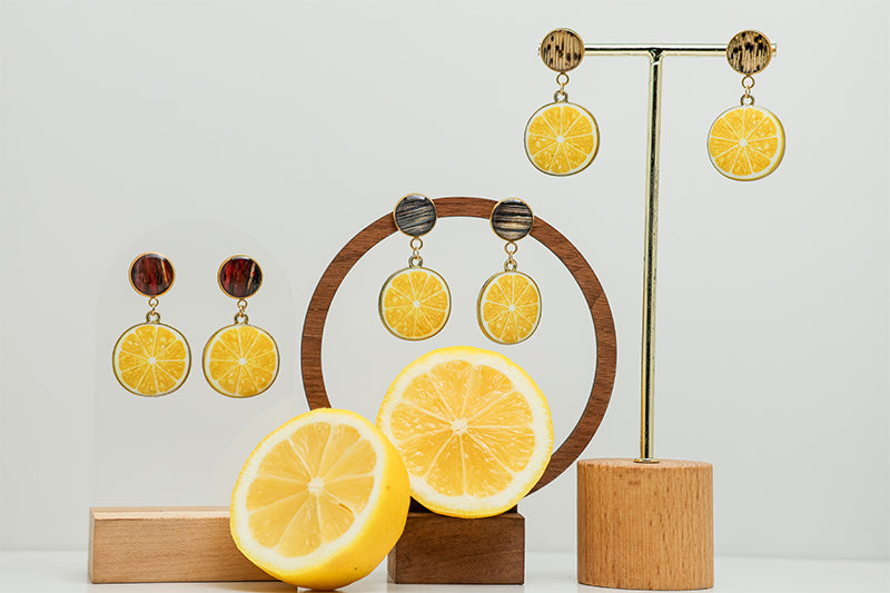 Three pairs of  Limoncello Earrings on stands on a white background with a lemon cut in half at the bottom of the image. 