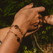 Hand wearing multiple gold bracelets and rings against a green leafy background