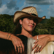 Woman wearing a cowboy hat and jewelry, standing next to a horse with a scenic background