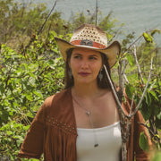 Woman wearing a cowboy hat and brown jacket standing in a natural setting with greenery and water in the background.