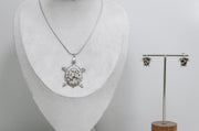Silver turtle pendant necklace on a mannequin bust and matching earrings on a stand against a gray background.