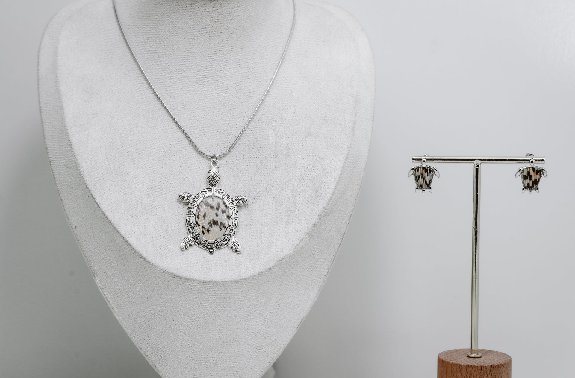 Silver turtle pendant necklace on a mannequin bust and matching earrings on a stand against a gray background.