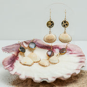 Small Scallop Shell Drops on a stand on a white background with two pairs in a pink clam shell at the bottom of the image with some sand. 