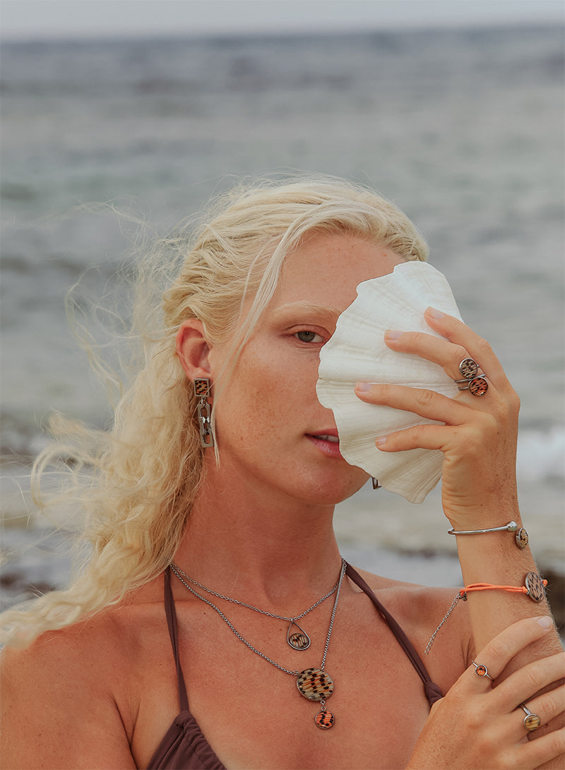 A woman in a brown bikini top in front of the ocean holding a conch shell wearing three rings, four bracelets, two necklaces and Chain Link Earrings.