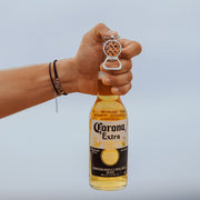 A hand with two bracelets holding a bottle of beer and a Silver Bottle Opener Keychain on a white background.