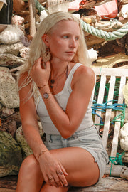 A woman sitting on a white bench in jean shorts and a white t-shirt wearing Wooden Mandala Statement Earrings, two bracelets and a necklace. 
