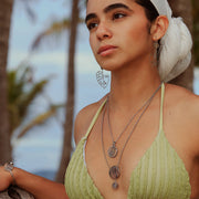 A woman in a light green bikini top and white hair scarf wearing two necklaces, a bracelet and earrings in front of palm trees.  