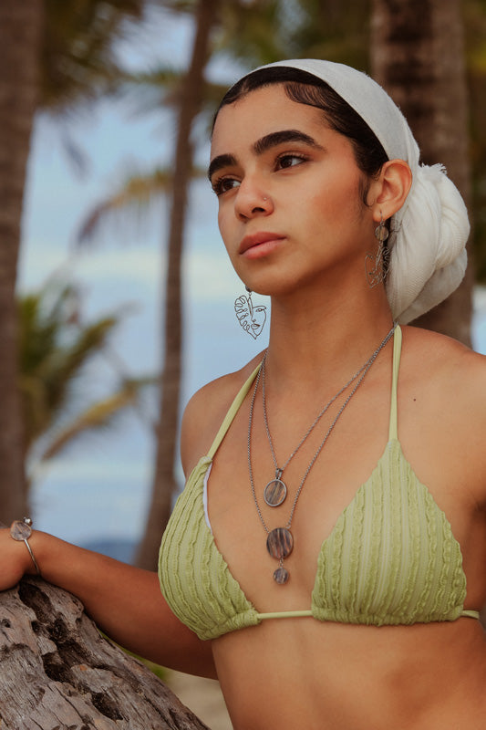 A woman in a light green bikini top and white hair scarf wearing two necklaces, a bracelet and earrings in front of palm trees.  