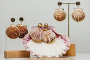 Three pairs of Scallop Shell Statement Earrings on stands on a white background with a pink clam shell at the bottom of the image.