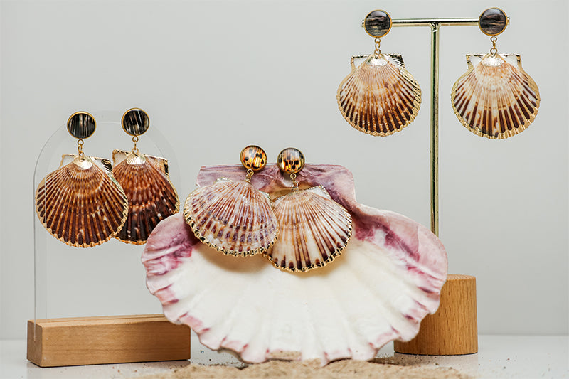 Three pairs of Scallop Shell Statement Earrings on stands on a white background with a pink clam shell at the bottom of the image.