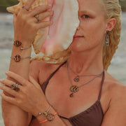 A woman in a brown bikini top in front of the ocean holding a conch shell wearing three rings, four bracelets, two necklaces and earrings. 
