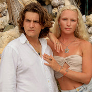 A man in a white shirt wearing. anecklace and a woman in jeans and a cream tube top wearing a necklace, two bracelets, three rings and earrings. They are standing in front of shells and rocks. 