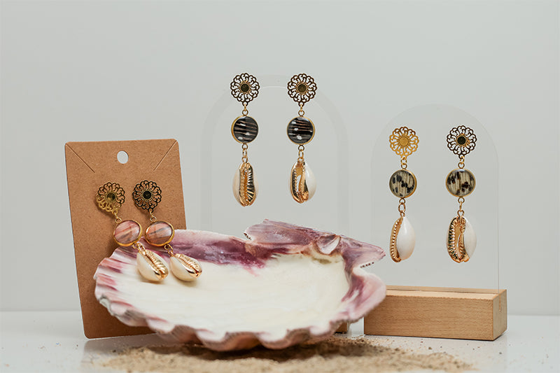 Three pairs of Cowrie Shell Mandala Drops on stands on a white background with a pink clam shell at the bottom of the image.