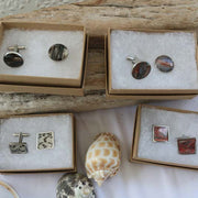 Four jewelry boxes with either a Round Cuff Links in the top row andSquare Cuffs in the bottom row. Link set in the box on a piece of wood on a white tablecloth with three shells on a white tablecloth.