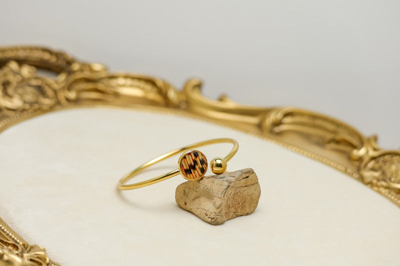 Gold cuff bracelet with lionfish tail fin resin pendant and gold ball end displayed on natural stone within ornate gold decorative tray