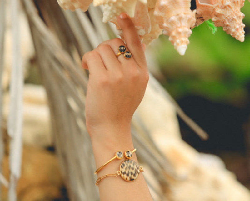 Gold paperclip bracelet with lionfish pendant, gold double pendant bracelet with two lionfish fin resin charms and gold double pendant ring worn on a wrist and hand with coastal shells and greenery background