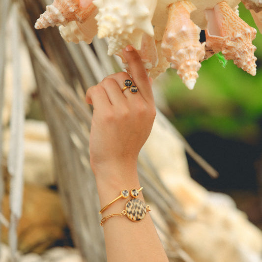 Two gold lionfish bracelets and a gold lionfish ring worn on model's wrist and hand with coastal shells and greenery background