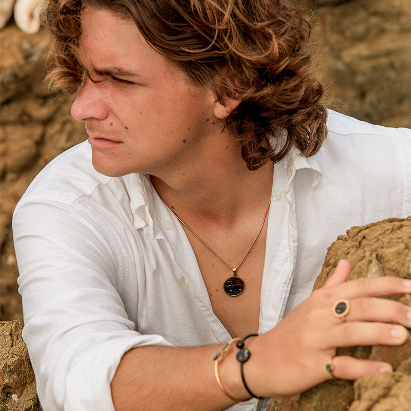 Man wearing gold lionfish fin pendant necklace with dainty chain, bracelet, and rings in open white linen shirt against rocky outdoor background