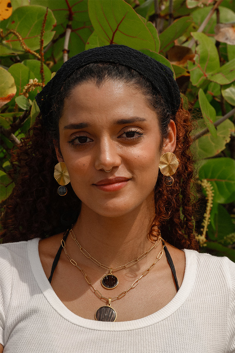 Woman wearing gold layered lionfish fin pendant necklaces and gold coin earrings in white t-shirt with green tropical foliage background