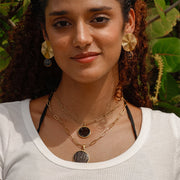 Woman wearing gold layered lionfish fin pendant necklaces and gold coin earrings in white t-shirt with green tropical foliage background