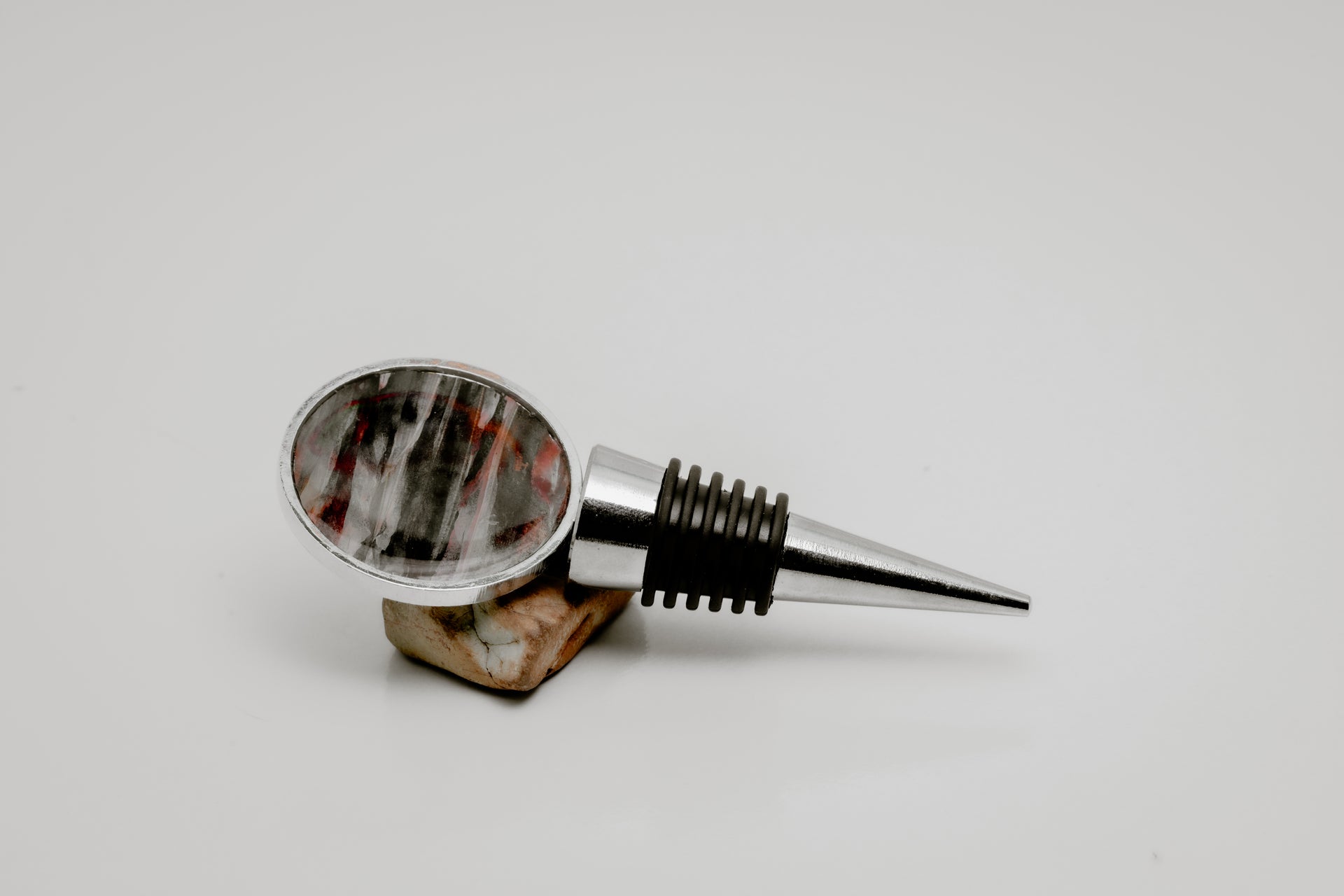 Silver wine bottle stopper with round lionfish pectoral fin resin charm in red and grey streaked pattern displayed on natural stone on white background
