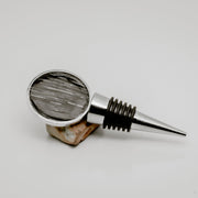 Silver wine bottle stopper with round lionfish pelvic fin resin charm in black and grey streaked pattern displayed on natural stone on white background