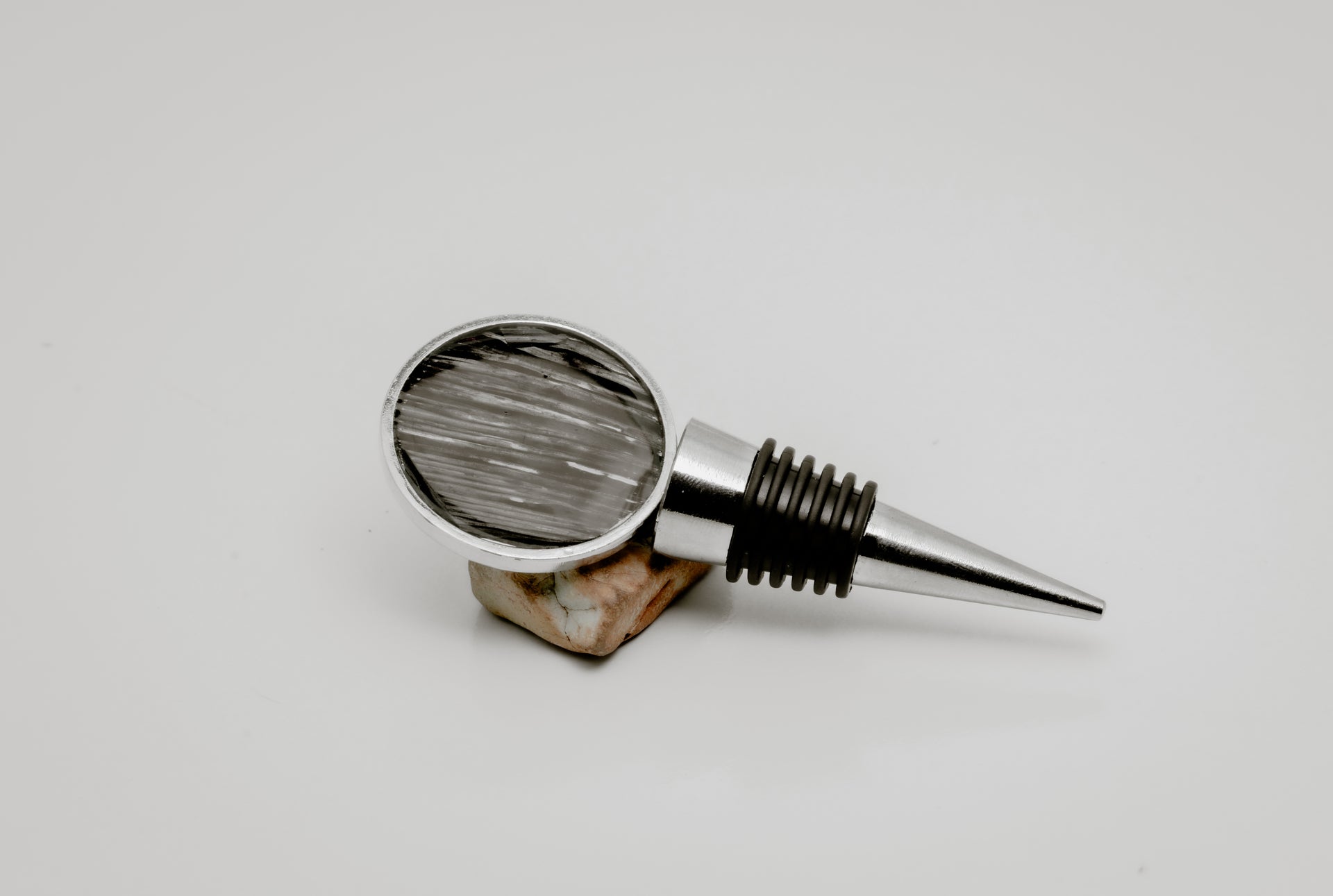 Silver wine bottle stopper with round lionfish pelvic fin resin charm in black and grey streaked pattern displayed on natural stone on white background